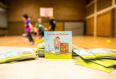 Ein Pixi Buch steht aufgestellt auf einem Tisch. Im Hintergrund sieht man Kinder,die in einer Sporthalle spielen.