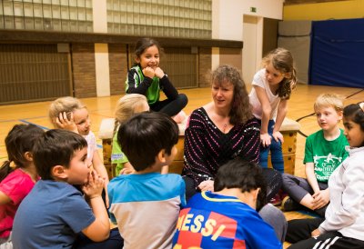 Staatssekretärin Andrea Milz sitzt umringt von Grundschulkindern in einer Sporthalle. IN ihrer Hand hält sie ein Pixi-Buch.