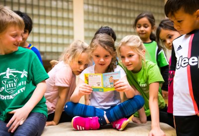 Eine Gruppe vo Grundschulkindern schauen sich ein Pixi-Buch an. Im Hintergrund sieht man die Glaskachelwand der Sporthalle.