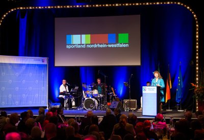 Staatssekretärin Andrea Milz am Rednerpult rechtsseitg auf einer Bühne. Links sitzt eine Musikband. Vor der Bühne sitzt das Publikum.