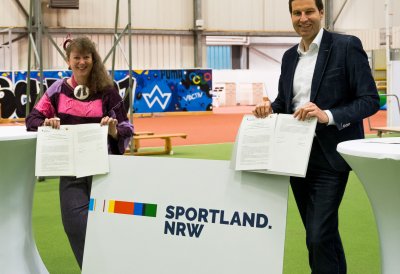Links im Bild steht Staatssekretärin Andrea Milz, sie  hat eine pink-schwarze Sportjacke an. Rechts im Bild steht ein Mann im schwarzen Anzug und weißem Hemd. Zwischen ihnen steht unten auf dem Boden ein  weißer Austeller mit dem "Sportland NRW"-Logo. Sie befindne sich in einer Indoor-Sporthalle.