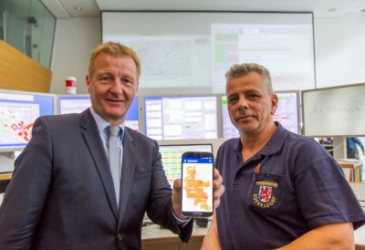 Innenminister Ralf Jäger zeigt auf dem Mobiltelefon neben einem Feuerwehrmann die Notfall-, Informations- und Nachrichten-App NINA 