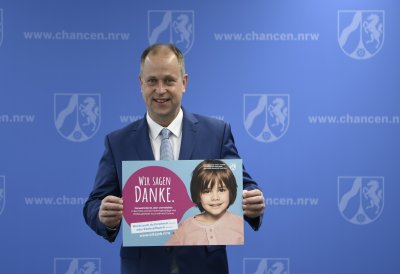 Ein Mann im Anzug steht vor einer blauen Wand und hält ein helblaues Plakat hoch, auf dem ein Kind zu sehen ist