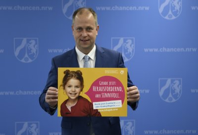 Ein Mann im Anzug steht vor einer blauen Wand und hält ein gelbes Plakat hoch, auf dem ein Kind zu sehen ist