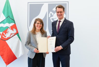 Rechts neben der NRW-Fahne steht Verena Schäffer mit langen blonden Haaren und Brille im grauen Sakko und weißem Hemd lächelnd neben MP Wüst im schwarzen Anzug mit roter Krawatte