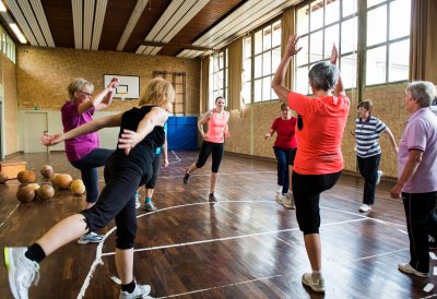Neun Damen in Sportbekleidung stehen in der Mitte einer Sporthallte im Kreis und machen Aufwärmübungen.