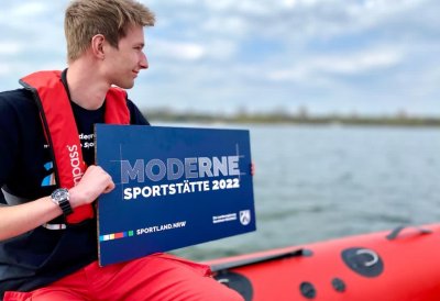 Ein junger Mann sitzt auf einem Schlauchboot und hält ein blaues Schild 