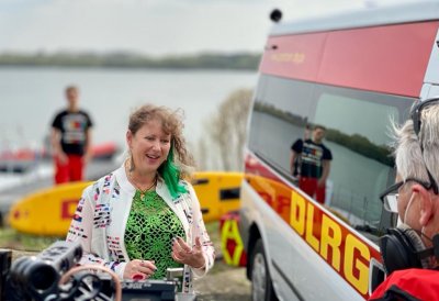 Eine Frau spricht vor einer Kamera, im Hintergrund ist ein Rettungswagen des DLRG