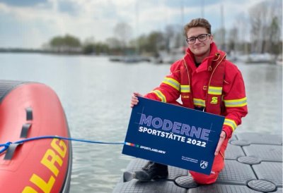 Ein Mann kniet auf einem Steg neben einem Schlauchboot und hält ein blaues Schild 