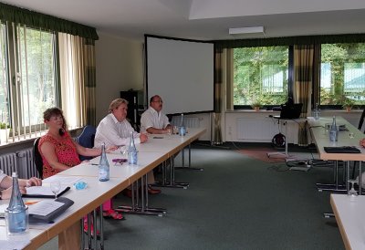Staatssekretärin Milz sitzt mit drei weiteren Herren an einer Tischreihe in einem Saal, gegenüber sitzt ein Herr im schwarzweiß-kariertem Hemd.