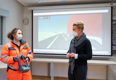 Eine Straßenwärterin im orangenen Overall zeigt der Ministerin eine VR-Brille vor einer Leinwand