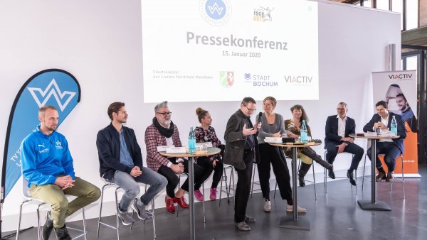 Staatssekretärin Andrea Milz sitzt mit Teilnehmern der Pressekonferenz auf Hockern an Rundtischen. Hinter ihnen eine weiße Rückwand mit einer Präsentation.