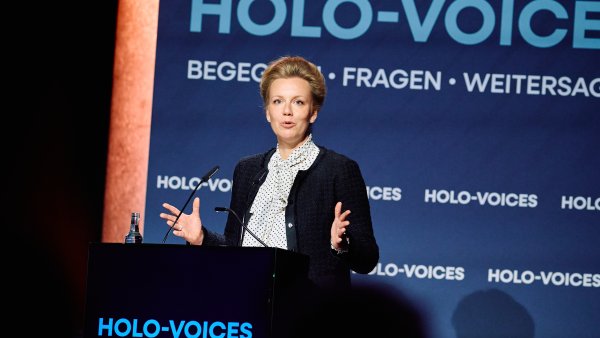 Eine Frau in dunklem Blazer steht gestikulierend an einem Redner-Pult 