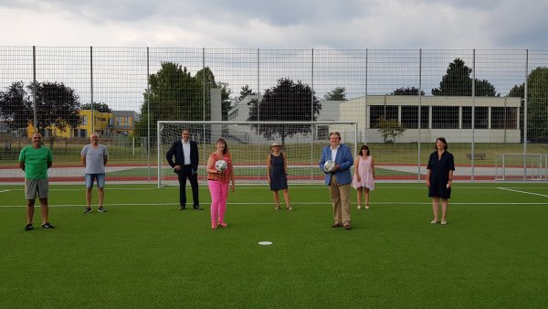 Staatssekretärin MIlz mit Vereinsmitgliedern vor dem Tor eines Rasen-Fußballplatzes. Sie in rosapinker Sportbekleidung mit einem Fußball in der Hand.