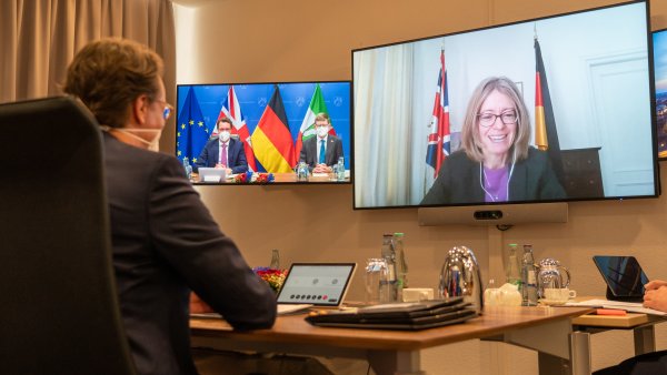 Ein Mann am Schreibtisch schaut auf zwei Bildschirme an der Wand. Im linken sind zwei Männer, im rechten eine Frau vor Flaggen zu sehen.