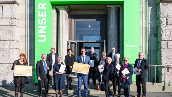 Eine Gruppe Menschen steht vor einem Eingang mit grünem Tor, zwei halten jeweils eine große Karte