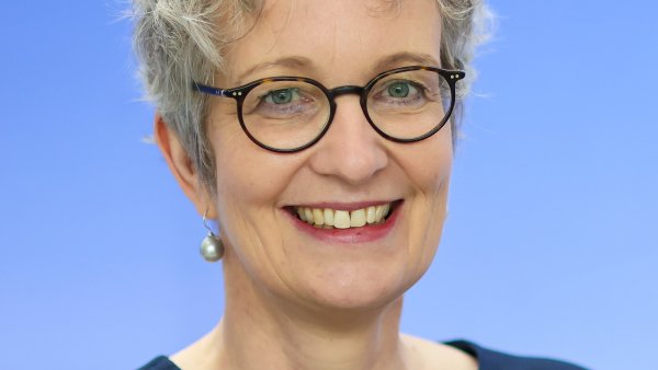 Eine Frau mit kurzem grauen Haar und Brille vor blauem Hintergrund