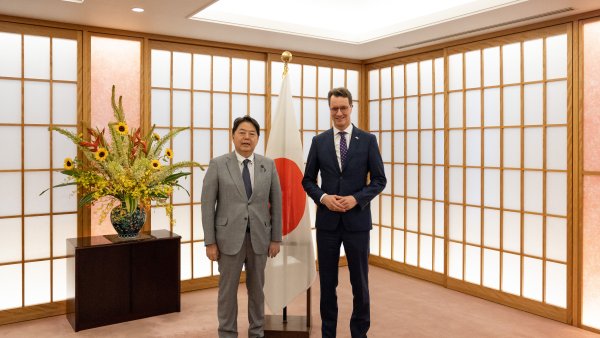 Zwei Männer in Anzügen stehen in einem japanischen Raum mit Japan-Flagge und einer Kommode und Blumen im Hintergrund