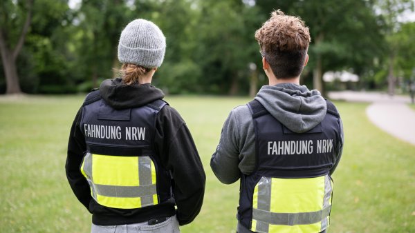 zwei Ermittler stehen mit schwarz-gelben Westen mit der Aufschrift "Fahndung NRW" vor einer Wiese in einem Park