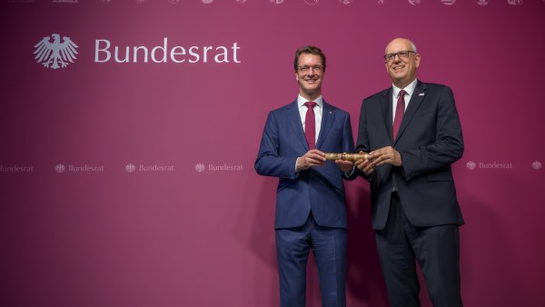 Zwei Männer in Anzügen halten gemeinsam einen Holzstab fest.