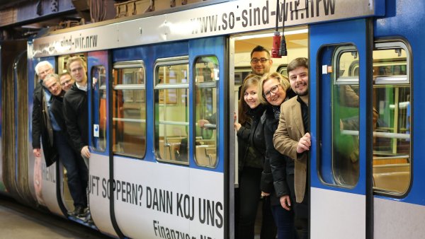 Ein Gruppe Menschen schaut aus einer U-Bahn, die mit der Werbung der Finanzverwaltung NRW verziert ist