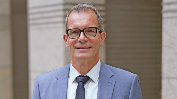 Ingo Dudenhausen steht im blauen Anzug und dunkelblauer Krawatt und weißem Hemd vor beigen Säulen. Er trägt eine Brille und lächelt frontal in die Kamera.