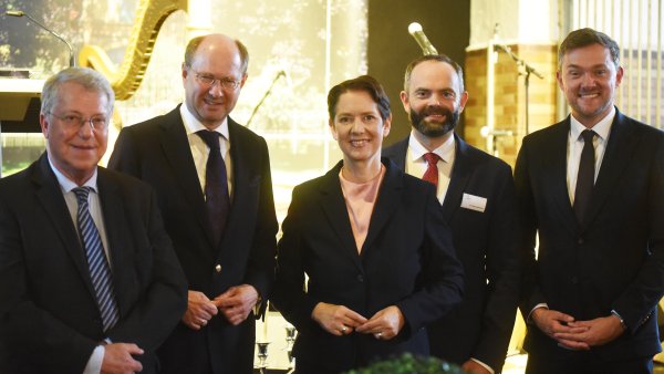Ministerin Silke Gorißen steht in der Mitte einer Gruppe aus fünf Personen und lächelt in die Kamera. Die vier Männer tragen Anzüge und Krawatten. Die Gruppe steht in einem hellen Innenraum vor großen Fenstern und einer Harfe.