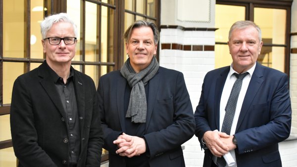 drei Männer stehen vor einer weißen Säule und hell beleuchtetem Hintergrund