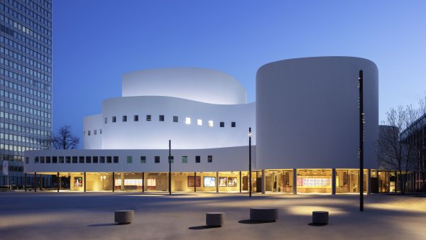 Das Düsseldorfer Schauspielhaus bei Dämmerung mit beleuchteter, geschwungener weißer Fassade und markanten Baukörpern. Im Vordergrund liegt ein weitläufiger Platz, flankiert von Hochhäusern.