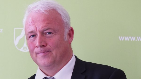 ein Mann mit grauem Haar, dunklem Sakko und gestreifter Krawatte steht vor einer grünen Wand