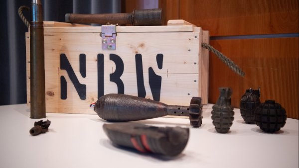 Holzkiste mit der Aufschrift „NBW“ und verschiedenen historischen Sprengkörpern und Granaten auf einem Ausstellungstisch. Die Objekte sind vor einem warm beleuchteten Hintergrund arrangiert und wirken wie Exponate in einer Ausstellung oder Sammlung.