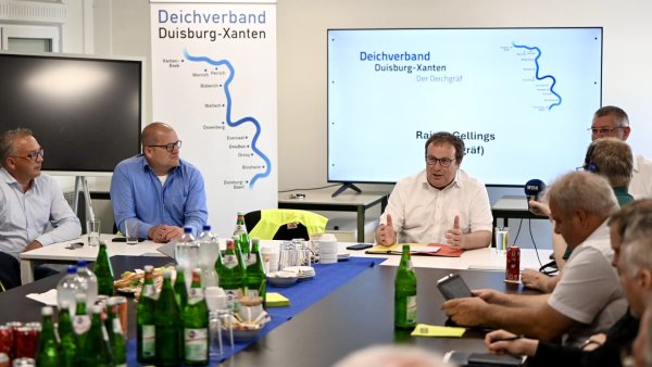 Umweltminister Oliver Krischer sitzt in einem weißen Hemd an einem Konferenztisch und spricht mit mehreren Personen. Im Hintergrund sind ein Bildschirm und ein Banner des Deichverbands Duisburg-Xanten zu sehen.