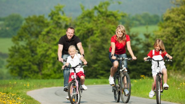 Eine Familie fährt mit Fahrrädern über einen Asphaltweg zwischen grünen Wiesen und Büschen. 