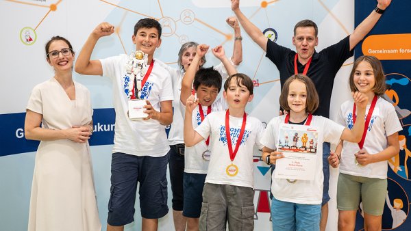 5 Kinder und 3 Erwachsene jubeln, die Kinder haben Medaillen an roten Bändern um den Hals