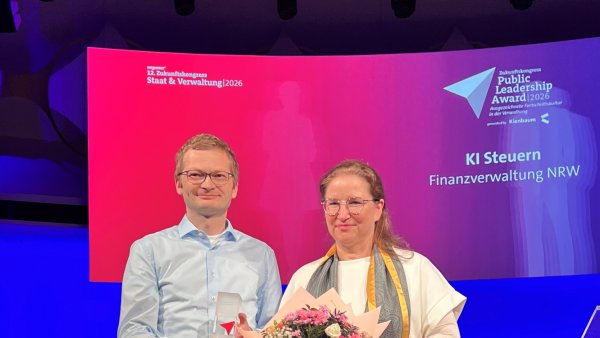 Zwei Personen stehen auf einer Bühne und präsentieren einen Preis sowie einen Blumenstrauß vor einer großen Leinwand. Im Hintergrund ist die Auszeichnung für das Projekt ‚KI Steuern‘ der Finanzverwaltung NRW beim Zukunftskongress Staat & Verwaltung 2026 zu sehen.