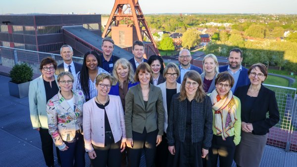 Gruppenfoto von etwa 15 Personen in formeller Kleidung auf einer Dachterrasse, die in die Kamera lächeln. Im Hintergrund steht ein großes Fördergerüst aus Stahl sowie Gebäude und eine grüne Landschaft. Die Personen stehen in zwei Reihen dicht beieinander.