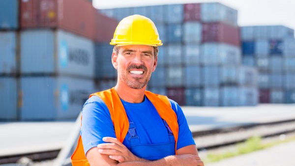 Ein lächelnder Hafenarbeiter mit blauem T-Shirt, orangefarbener Sicherheitsweste und gelbem Sicherheitshelm steht mit verschänkten Armen vor ein Paar Schienen. Im Hintergrund stehen mehrere Wände voller Container.