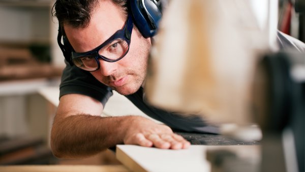 Ein Mann im schwarzen T-Shirt mit schwarzer Brille und Kopfhörern als Lärmschutz sägt ein Holzbrett an einer Maschine.