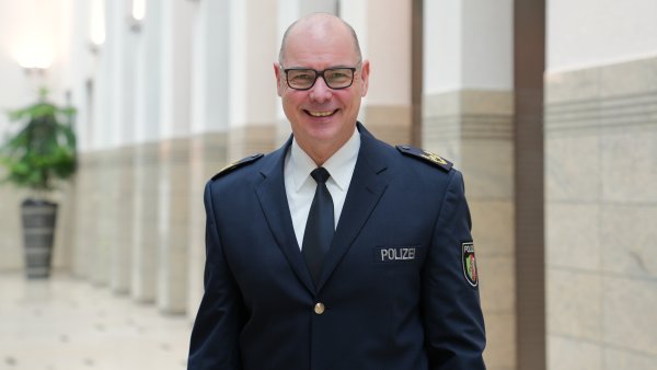 Dirk Hulverscheidt, der neue Inspekteur der Polizei NRW steht in blauem Polizeianzug lächelnd in einer Halle. Auf dem Bild ist nur sein Oberkörper zu sehen mit dunkelblauem Sakko, weißem Hemd und blauer Krawatte. Er trägt eine Brille und hat sehr kurz geschnittene Haare.
