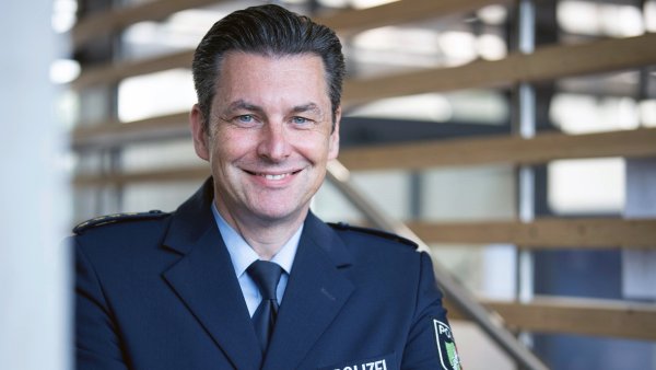 Markus Hattwig steht mit blauer Polizeiuniform (blaues Sakko mit Aufdruck POLIZEI, weißes Hemd, dunkelblaue Krawatte) lächelnd mit verschränkten Armen vor einer Wand aus Holzpaneelen