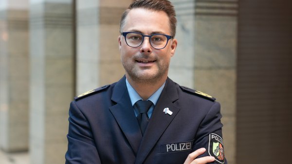 Ein Mann mit kurzen Haaren und Brille in Polizeiuniform und verschränkten Armen schaut lächeld in die Kamera
