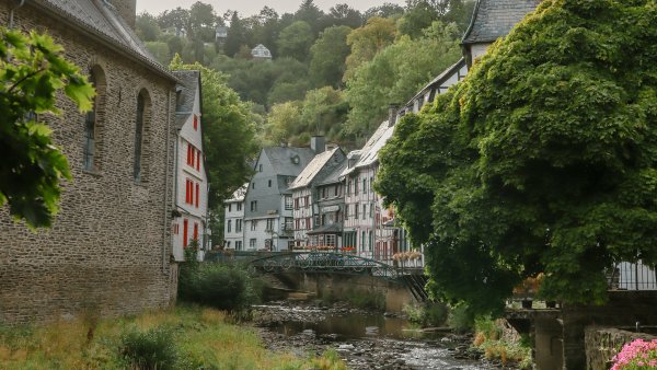 Historische Altstadt mit steinerner Kirche, Fachwerkhäusern und einem Bachlauf, eingerahmt von dichtem grünen Laub. Die idyllische Szene zeigt eine ruhige Flusslandschaft mit Brücke und blühenden Pflanzen.