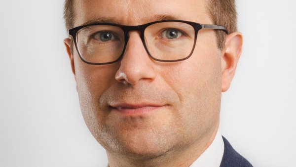 Ein Mann mit kurzen Haaren, Brille, weißen Hemd und blauem Jackett schaut in die Kamera