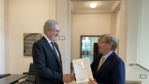 Zwei Männer stehen sich händeschüttelnd gegenüber, der rechte hält ein Dokument in der Hand