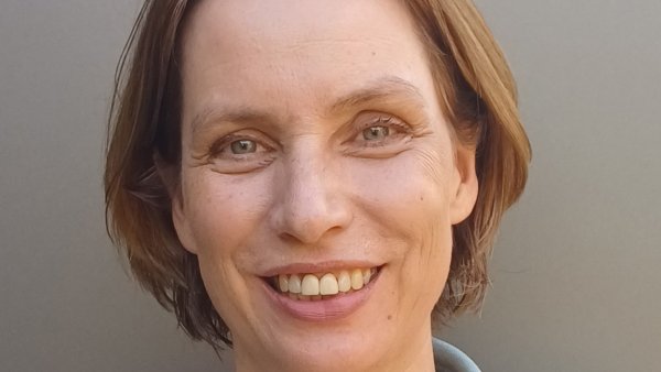Porträt einer lächelnden Frau mit kurzem, braunem Haar und freundlichem Blick. Sie trägt einen hellblauen Blazer über einem weißen Oberteil und blickt direkt in die Kamera. Der neutrale graue Hintergrund unterstreicht die professionelle und sympathische Ausstrahlung.