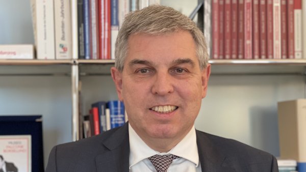 ein grauhaariger Mann mit Anzug und Krawatte vor einem Bücherregal