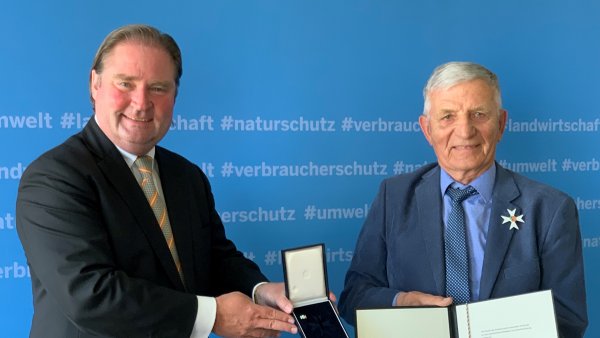 Zwei Männer in Anzügen stehen vor einer blauen Wand und halten einen Orden und ein Dokument