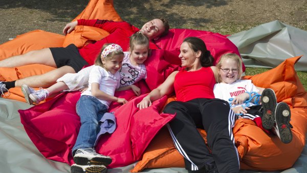 Mehrere Kinder ruhen sich mit ihrer Trainerin nach dem Sport auf großen, roten und orangefarbenen Kissen aus.