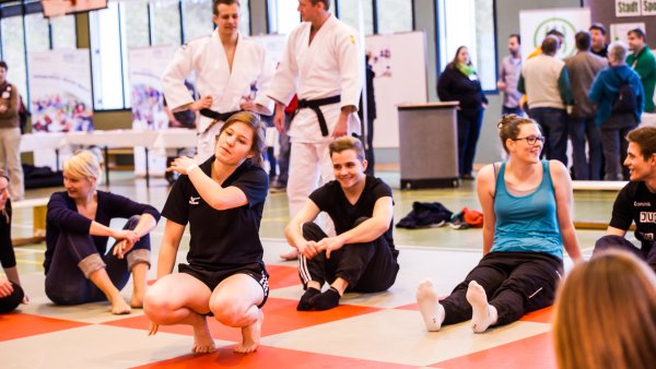 Mehrere Frauen bei Dehnübungen für einen Selbstverteidungskurs in einer Turnhalle. Auf dem Boden grüne und rote Matten im Schachbrettmuster. Im Hintergrund zwei Kampfsportler in weißen Anzügen mit schwarzen Gürteln.