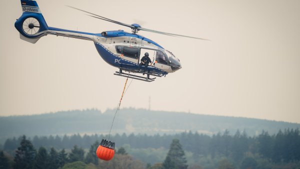 Zu sehen ist ein Löschhubschrauber der Polizei vor einem verrauchten Waldgebiet